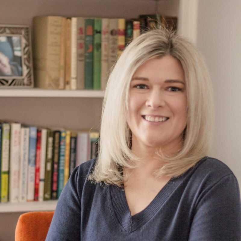 Emily Dein, a smiling blonde woman sitting in front of a book shelf