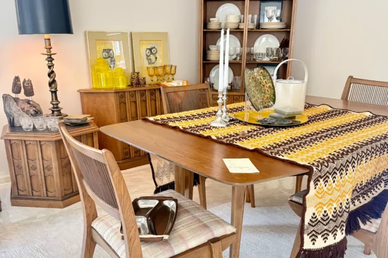 A vintage dining room setup featuring a wooden table covered by a thick yellow and brown crochet runner. Cabinets hold dishware, glassware, and owl-themed decor.
