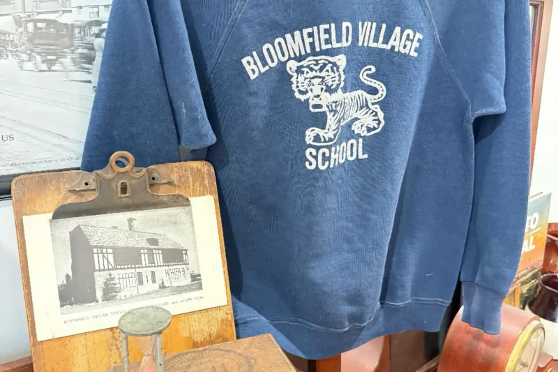 A vintage blue sweatshirt displaying a white tiger mascot graphic for "Bloomfield Village School." A wooden clipboard with an old photo of the school is in the foreground.