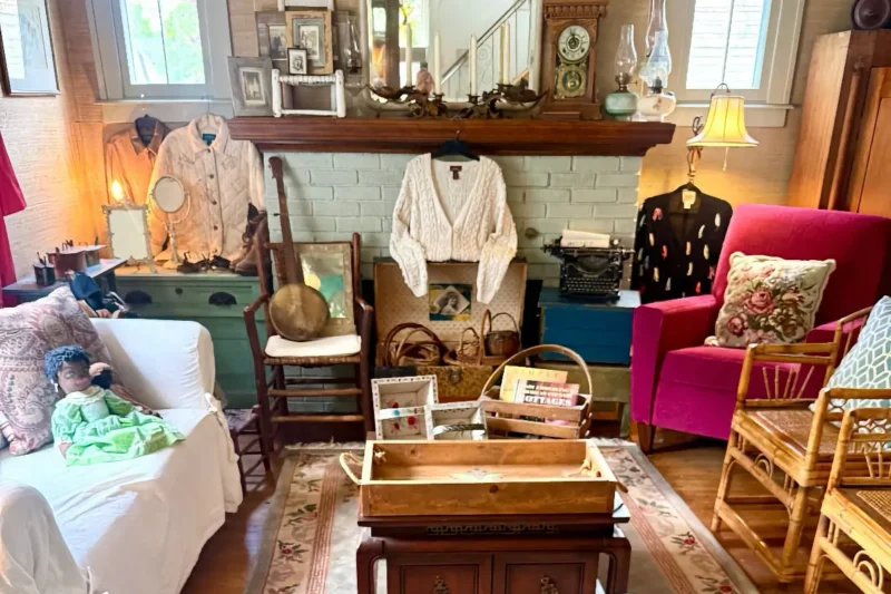 A densely furnished estate sale room displaying numerous vintage items like clothing, a clock, a pink velvet chair, baskets, and wooden furniture.