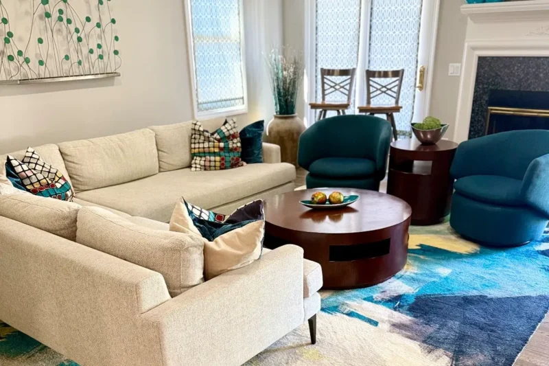 A contemporary living room featuring a neutral sectional sofa, round wood coffee table, and two dark teal swivel chairs over an abstract blue rug.